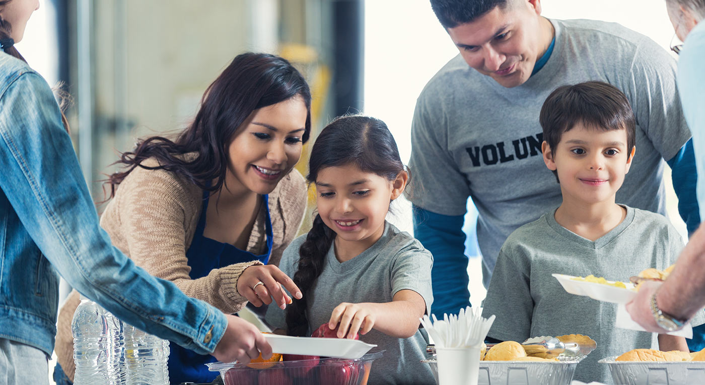Young Hispanic family is serving healthy meals to people in charity food bank soup kitchen. Mother and father are instructing elementary age little boy and little girl as they serve food to senior man and preteen girl.