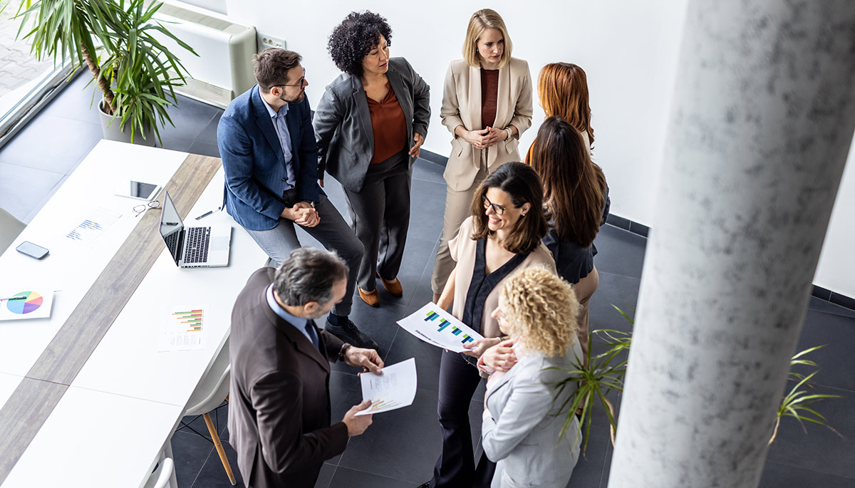 A group of professionals interactively discussing their projects and insights in a bright, modern office surrounding a large table with charts.