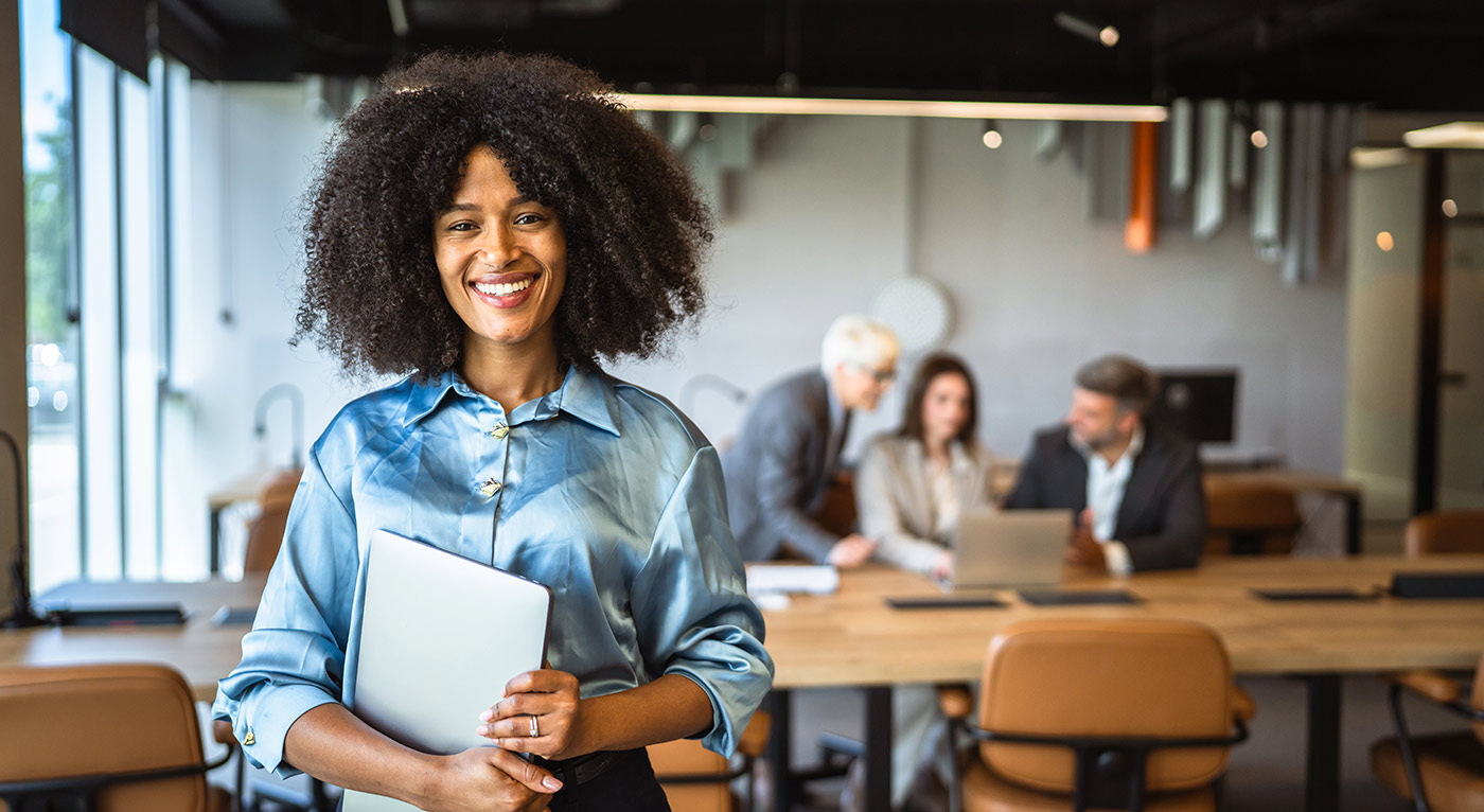 Instilling Knowledge and Confidence: Why Rising Generation Family Members Need Both Financial Acumen and Personal Empowerment A woman in a light blue blouse holding a laptop in an office, with colleagues in the background.