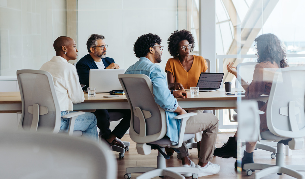 A medium-wide, eye-level shot captures five people in a conference room. The room has a large table, and the people are seated in ergonomic office chairs, with some using laptops.