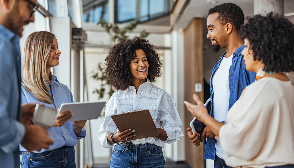 A diverse group of five casually dressed professionals, two men and three women, are standing and smiling while conversing in a bright office hallway.