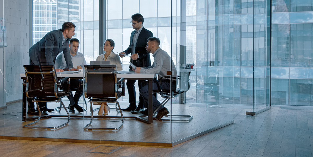 A group of six people having a meeting in a glass conference room with cityscape views.