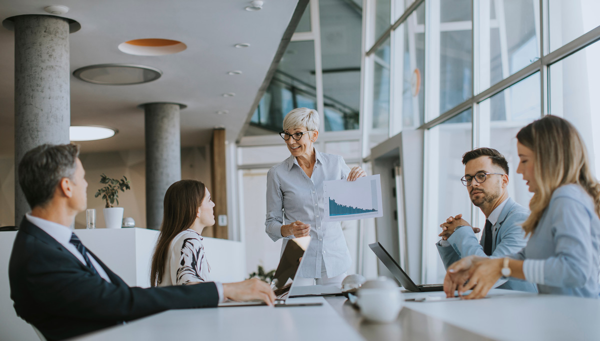 A woman presents a bar chart to four colleagues around a conference table in a modern office with large windows.