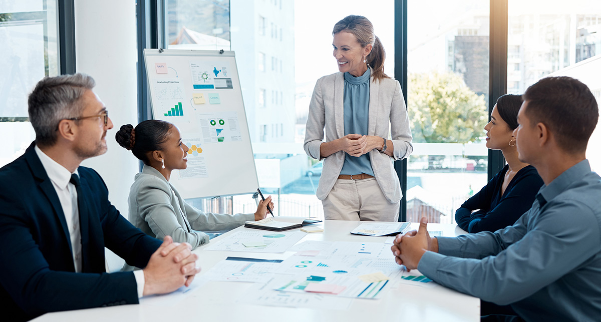 A professional woman gives a presentation to a seated group using a whiteboard with various charts and graphs.