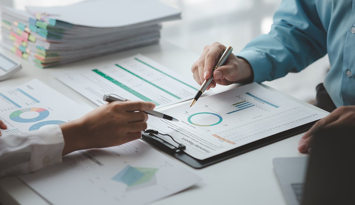 Close-up of hands using pens to point at data and colorful donut charts on a financial report document.