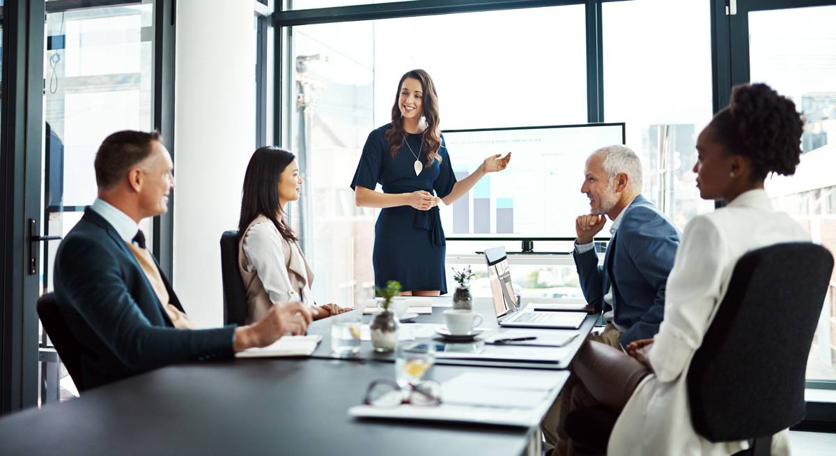 A professional woman standing at the head of a conference table delivers a presentation using a screen with bar charts.