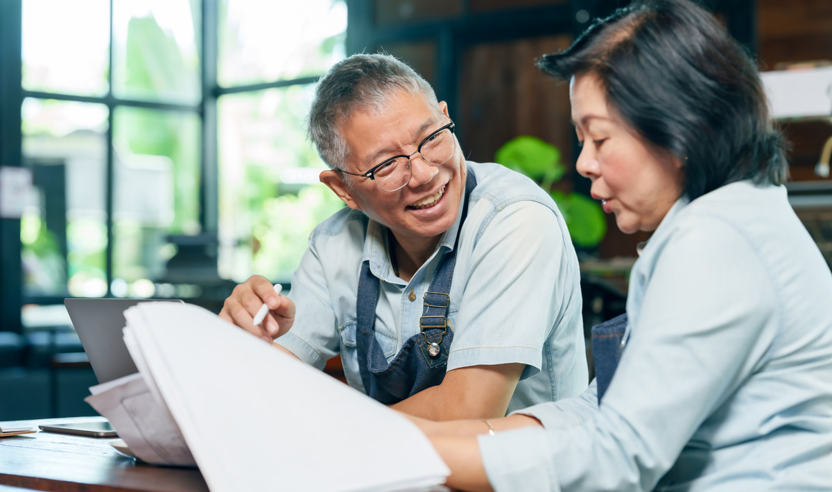 Senior business owners in aprons smile while reviewing paperwork and a laptop together in a bright, modern workspace.