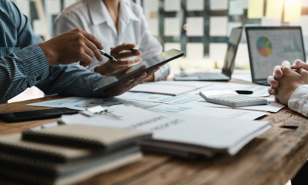Colleagues collaborate at a wooden desk, using a tablet, calculator, and laptops to review financial charts and reports in a modern office.