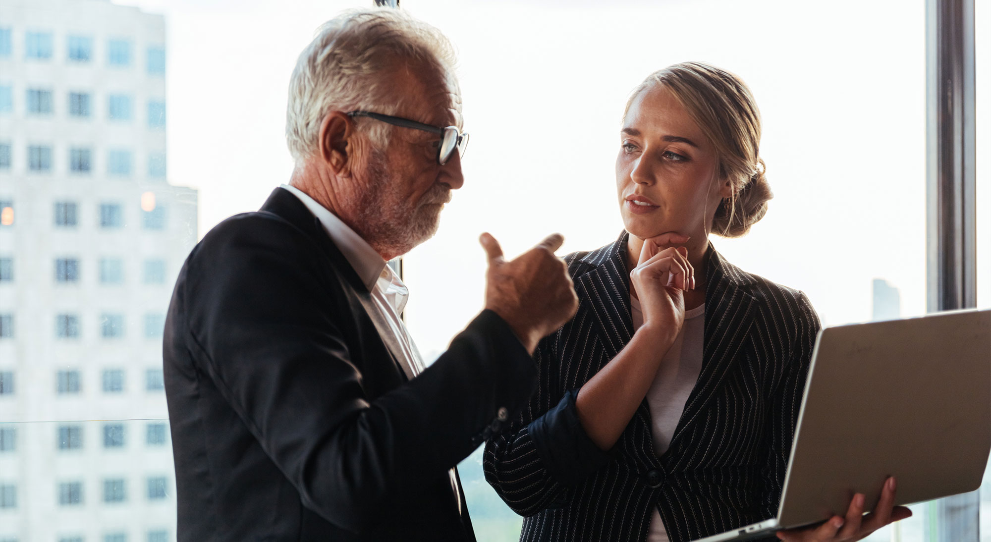 An older man in a suit gestures while speaking to a younger woman holding a laptop in front of a bright city window.