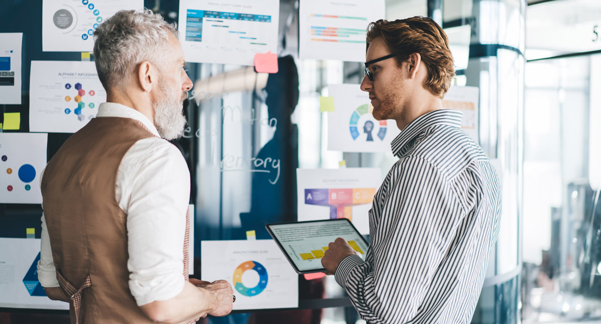 Two men in an office discuss data while standing in front of a glass wall covered in charts, graphs, and infographics.