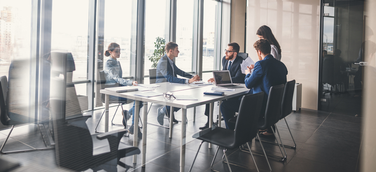 A business team in professional attire having a meeting around a conference table inside a bright, glass-walled office.