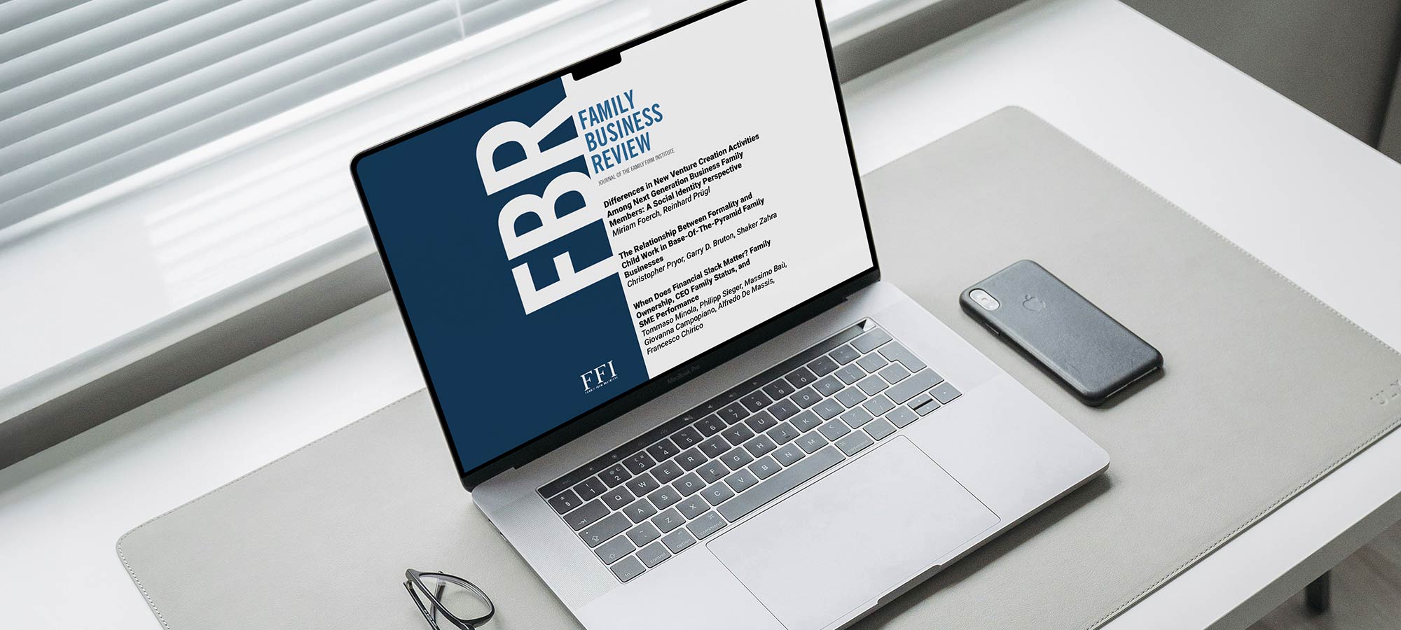 Laptop on a desk displaying the Family Business Review (FBR) journal cover from FFI. A smartphone and glasses sit on a grey desk mat next to the computer.