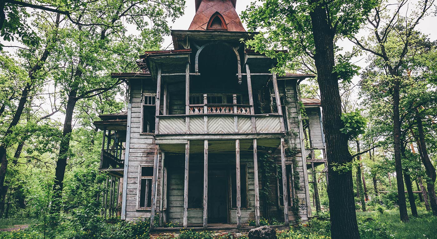 Abandoned wooden house surrounded by dense green trees, featuring a porch and a pointed tower.