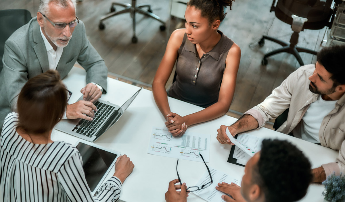 A group of people in a meeting around a table with a laptop and documents.