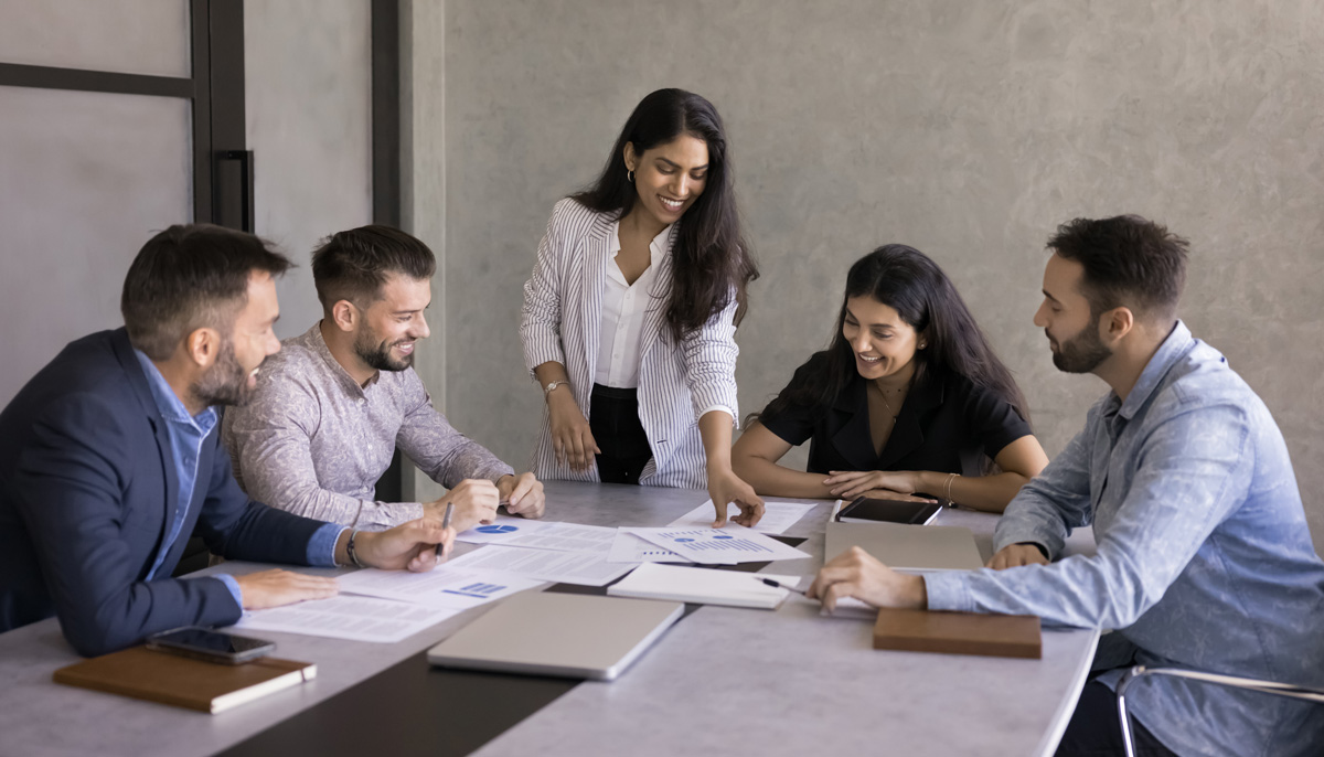 A smiling woman in a striped blazer stands to present documents to a seated group of colleagues collaborating around a conference table.