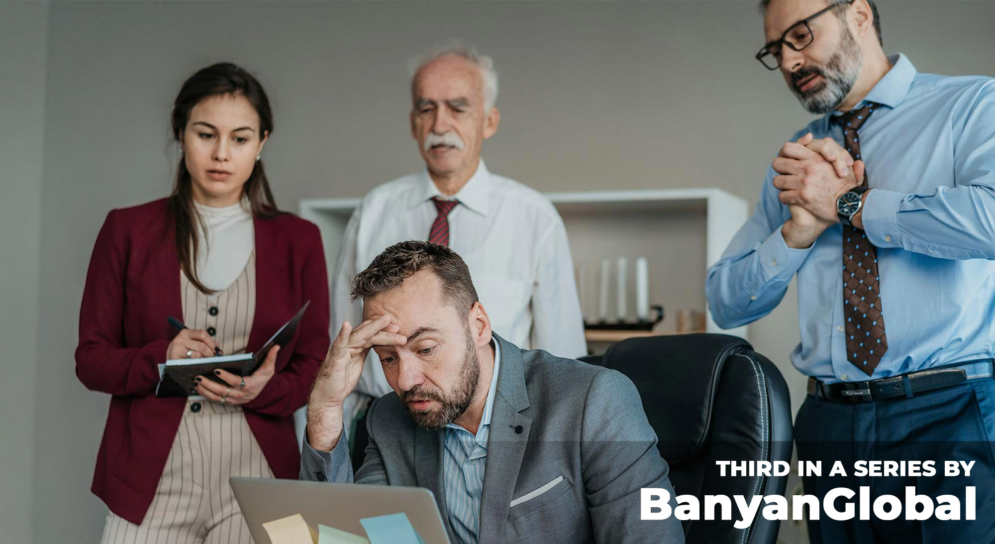 A stressed man looks down at a laptop while three family members surround him with concerned expressions.