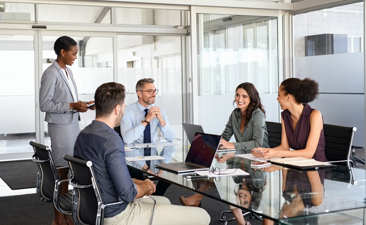 A woman stands presenting to four colleagues seated around a glass conference table in a modern office meeting room.