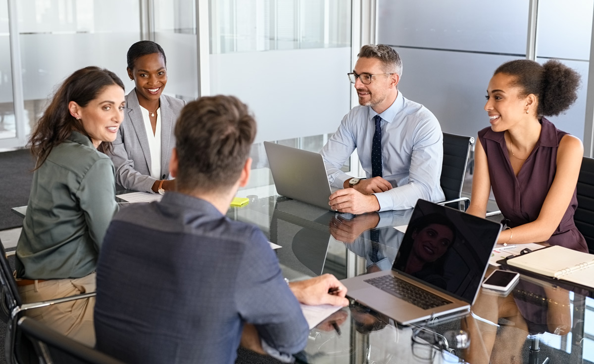A diverse group of five business professionals smiles and talks together around a glass conference room table.
