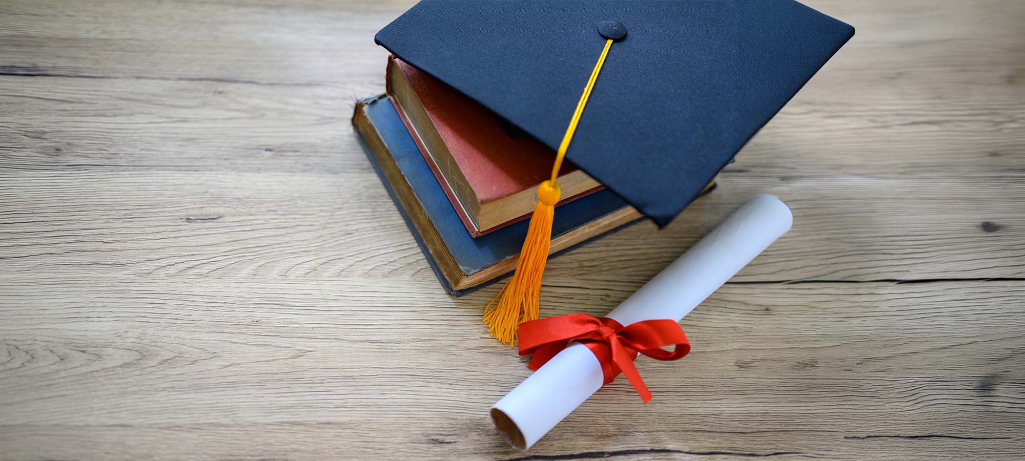 A graduation cap resting on a stack of books next to a tied diploma.