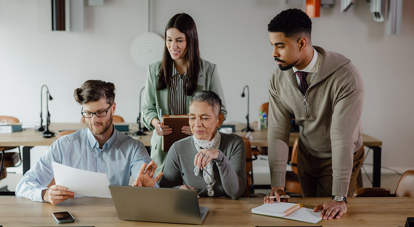 A diverse team gathers around a desk, collaborating over a laptop and printed documents.