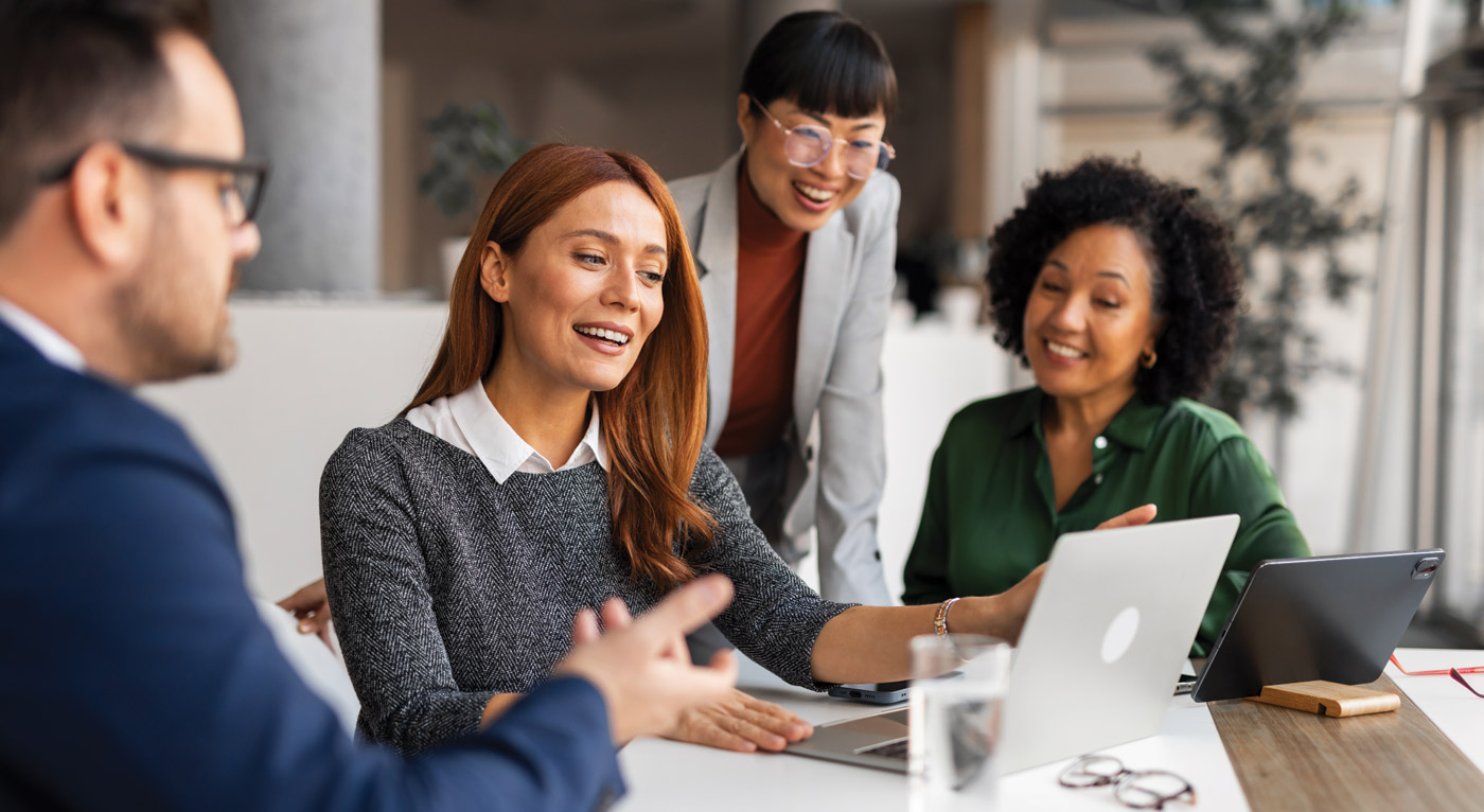 Group of four diverse professionals gathered around a laptop computer discussing a project together in a bright office environment.