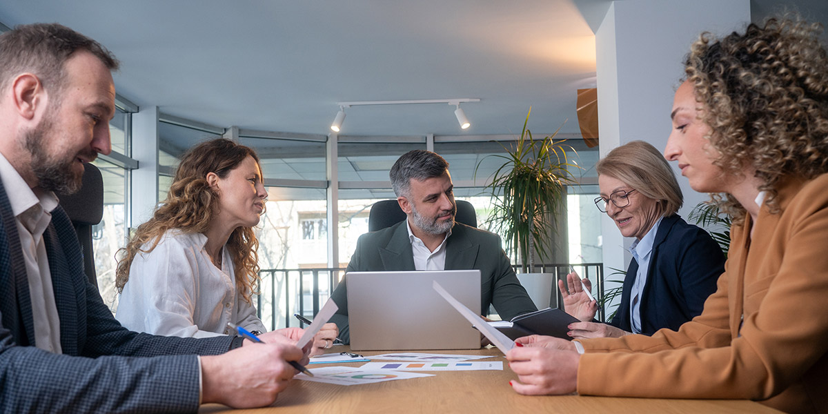 Five business professionals discussing documents around a boardroom table in a bright office.