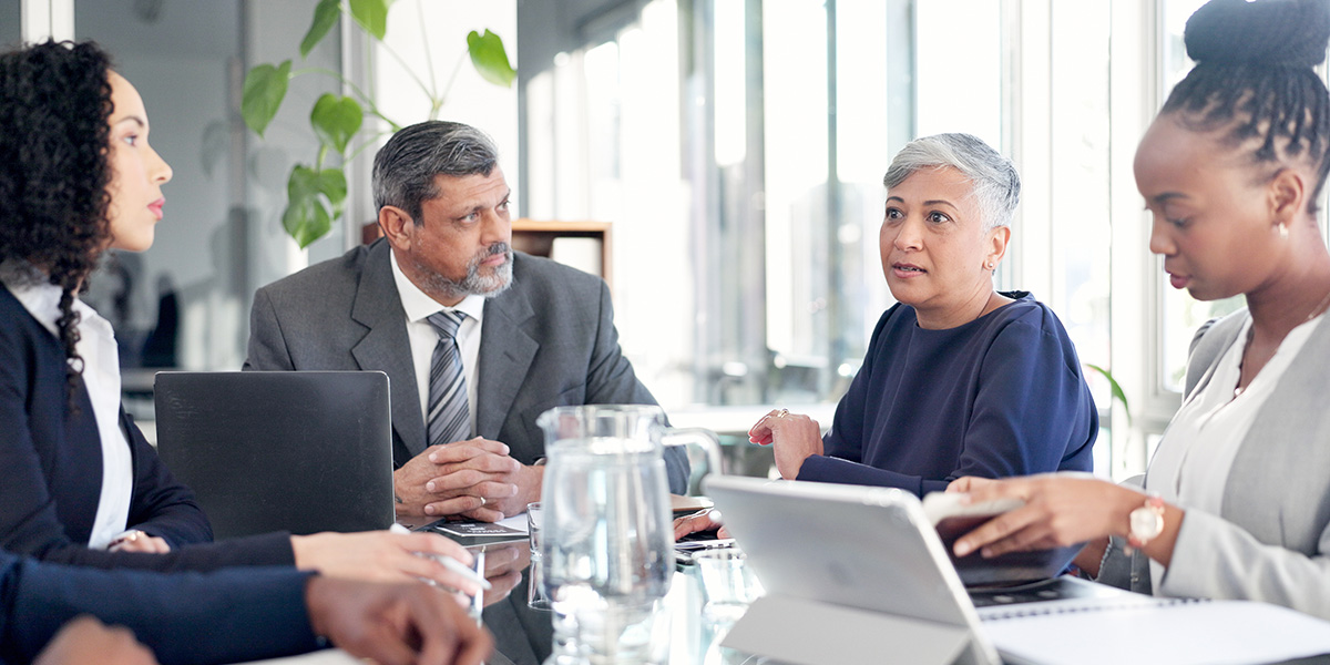 A diverse group of corporate executives seated at a glass conference table during a formal meeting, with a senior woman speaking.   