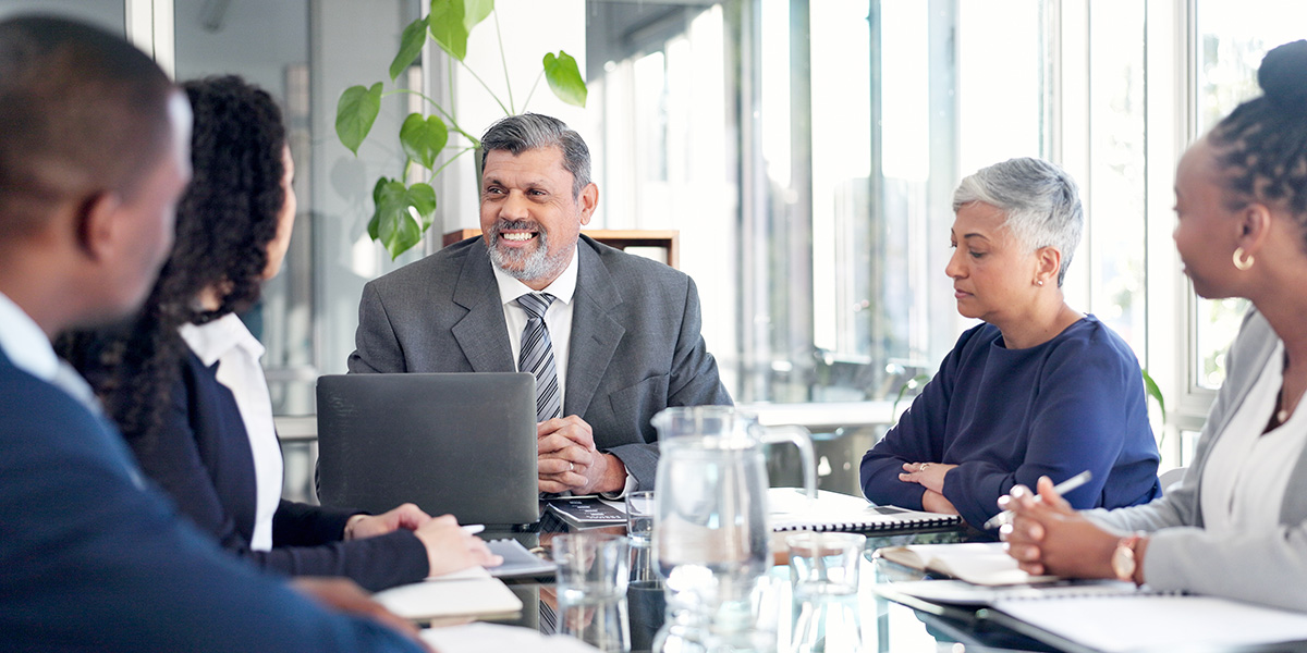 A professional team meeting at a glass table where a smiling male executive speaks to his seated colleagues.