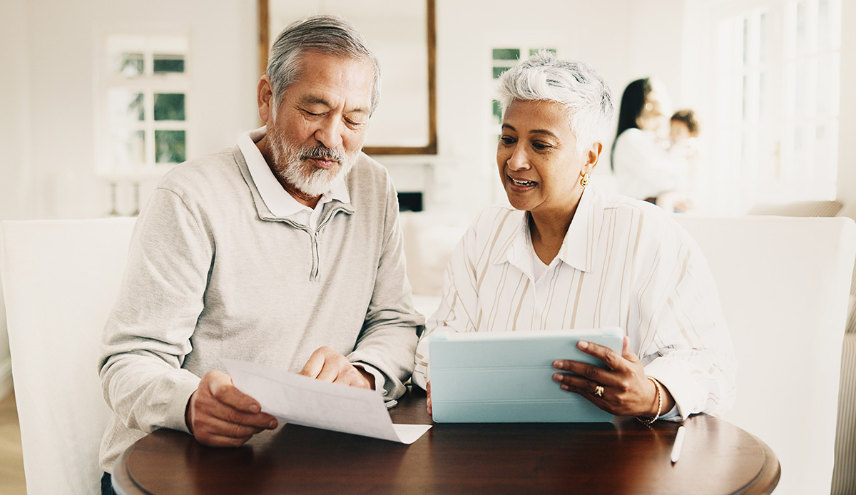 Senior couple sitting together at a table review a document while using a digital tablet.